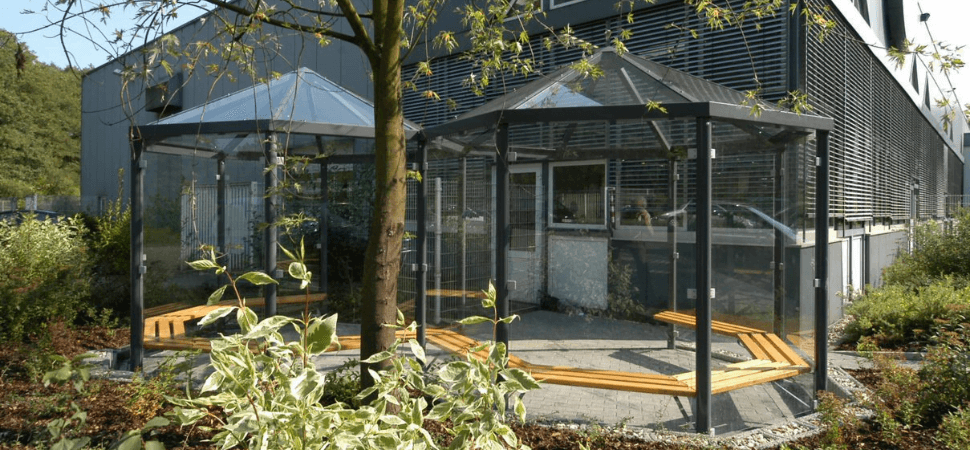 Buiten rookruimte met glazen wanden en zitbanken, dubbele overkapping in stalen frame, geplaatst in groene bedrijfsomgeving.