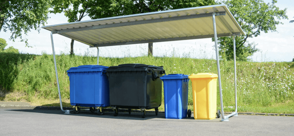 Materiaaloverkapping voor afvalbakken en rolcontainers, geplaatst op asfalt langs een groene strook.