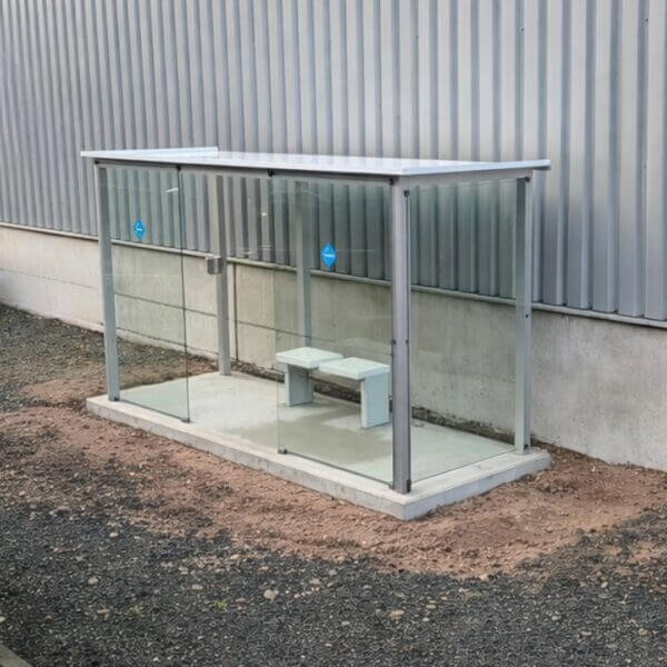 Kant en klare rookabri kopen Moderne rookabri van glas en geanodiseerd aluminium met zitbank en asbak, professioneel geplaatst op een betonplaat naast een bedrijfspand.