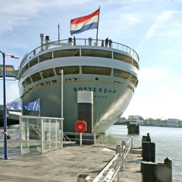 Rookcabine bij SS Rotterdam Grote cruisescheepsboeg van de SS Rotterdam met een Nederlandse vlag, naast een glazen rookcabine op de kade.