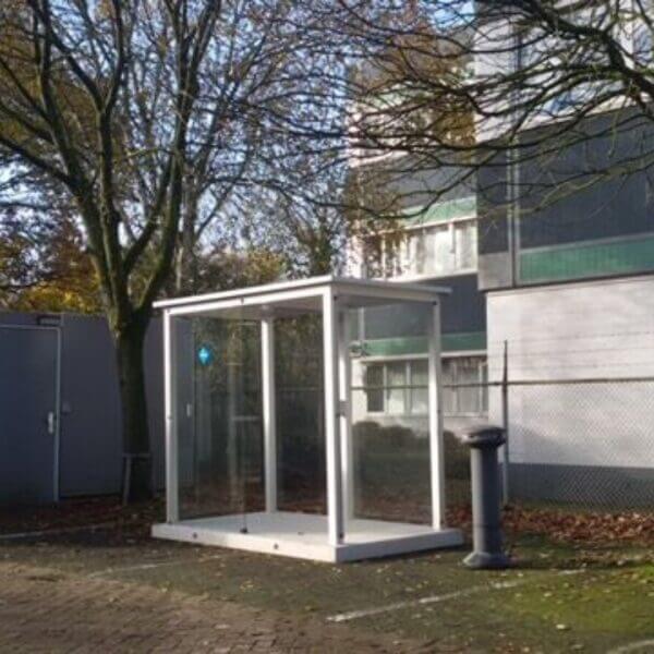 Compacte rookcabine voor buiten direct online kopen Prefab rookcabine van glas en aluminium, geplaatst op een prefab betonplaat, naast een kantooromgeving met bomen op de achtergrond.