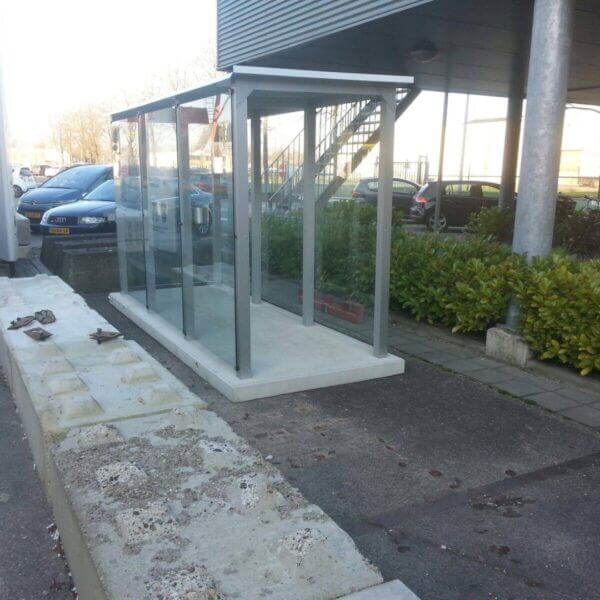 Prefab rookoverkapping op betonplaat Prefab rookoverkapping van glas en metaal op een prefab betonplaat, geplaatst naast een gebouw met parkeervakken.