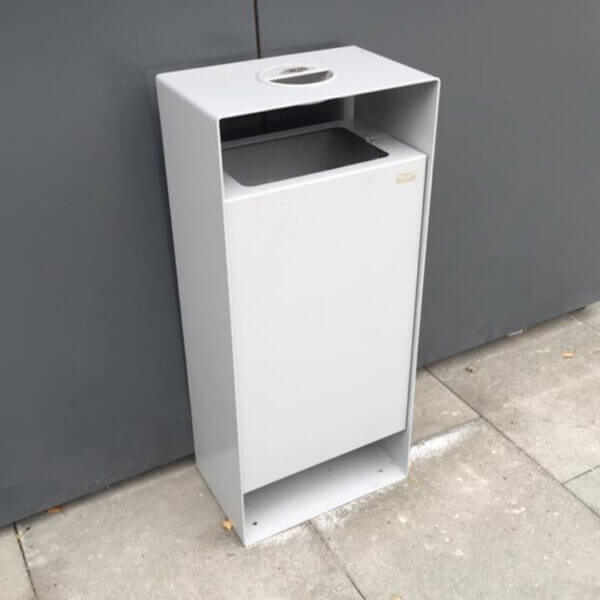 Prullenbak buiten 90 liter direct online bestellen Grijze buitenafvalbak van 90 liter met open bovenkant en asbak, geplaatst tegen een grijze muur.