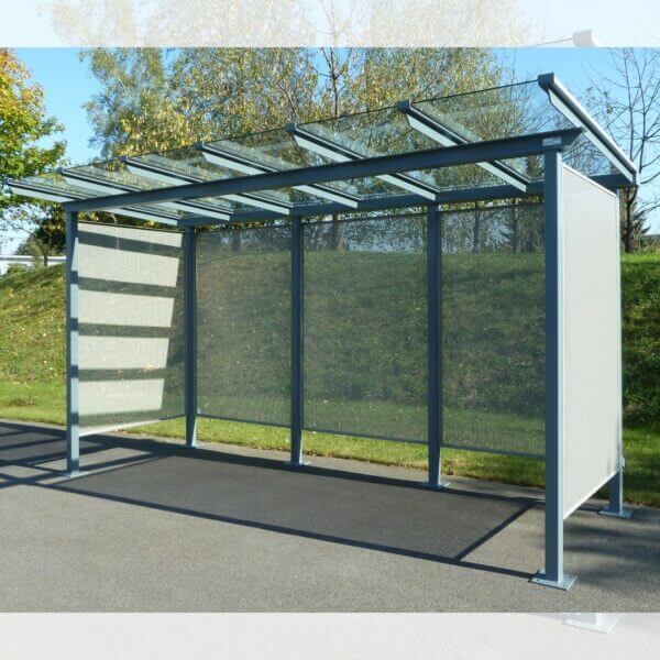 Multifunctionele fietsoverkapping met stalen wanden aanschaffen Fietsoverkapping met geperforeerde stalen zij- en achterwanden, dak van gehard glas in RAL-7016, verankerd op asfalt.