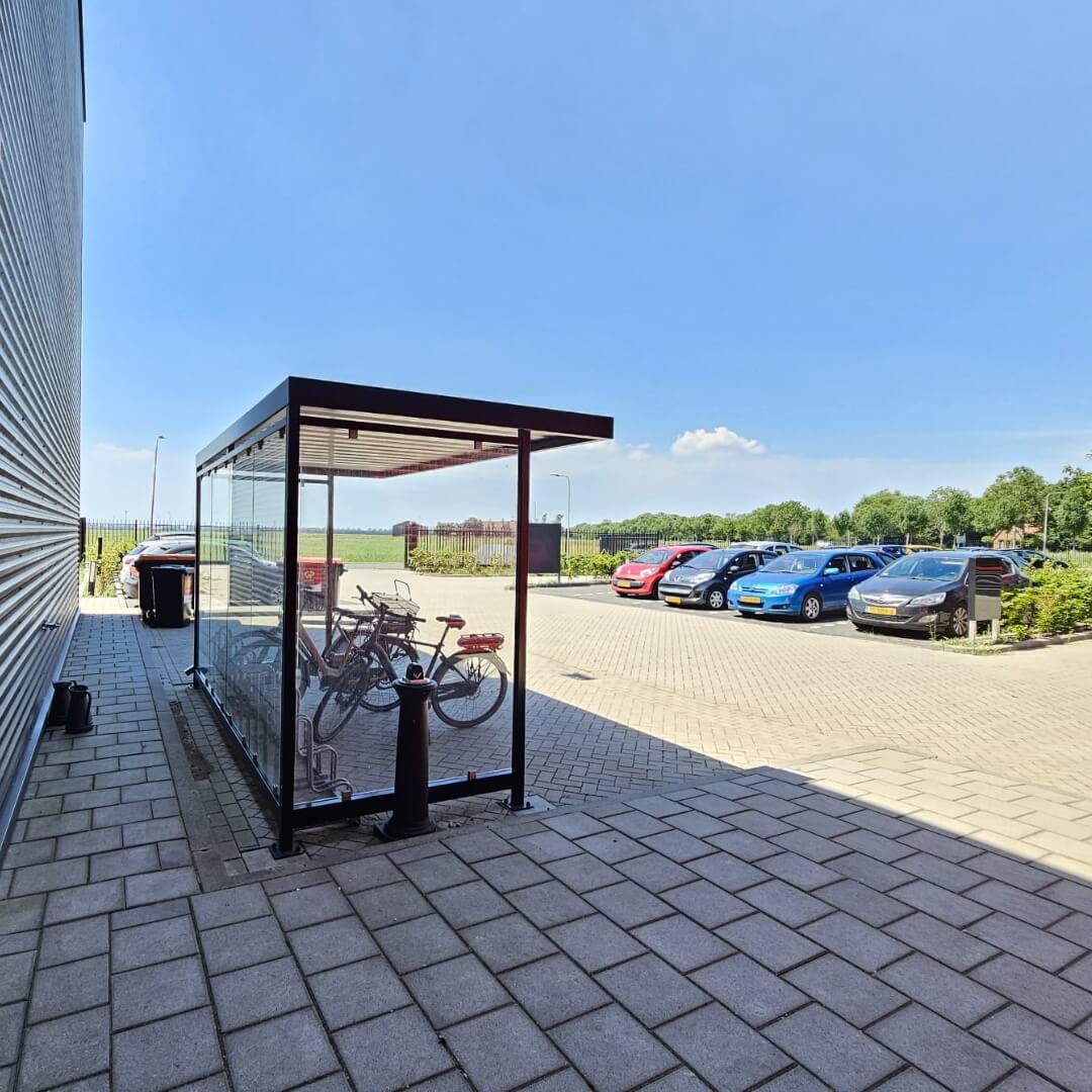Glazen fietsoverkapping met metalen frame, geplaatst langs een bedrijfsgebouw. Binnenin staan meerdere fietsen geparkeerd in een fietsenrek. Op de achtergrond is een parkeerplaats met auto's zichtbaar.
