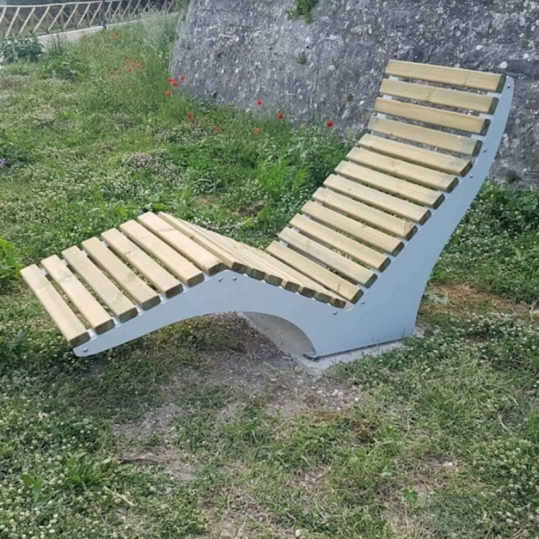 Ligstoel van hout en staal voor de openbare ruimte, geplaatst op een grasveld tegen een stenen muur