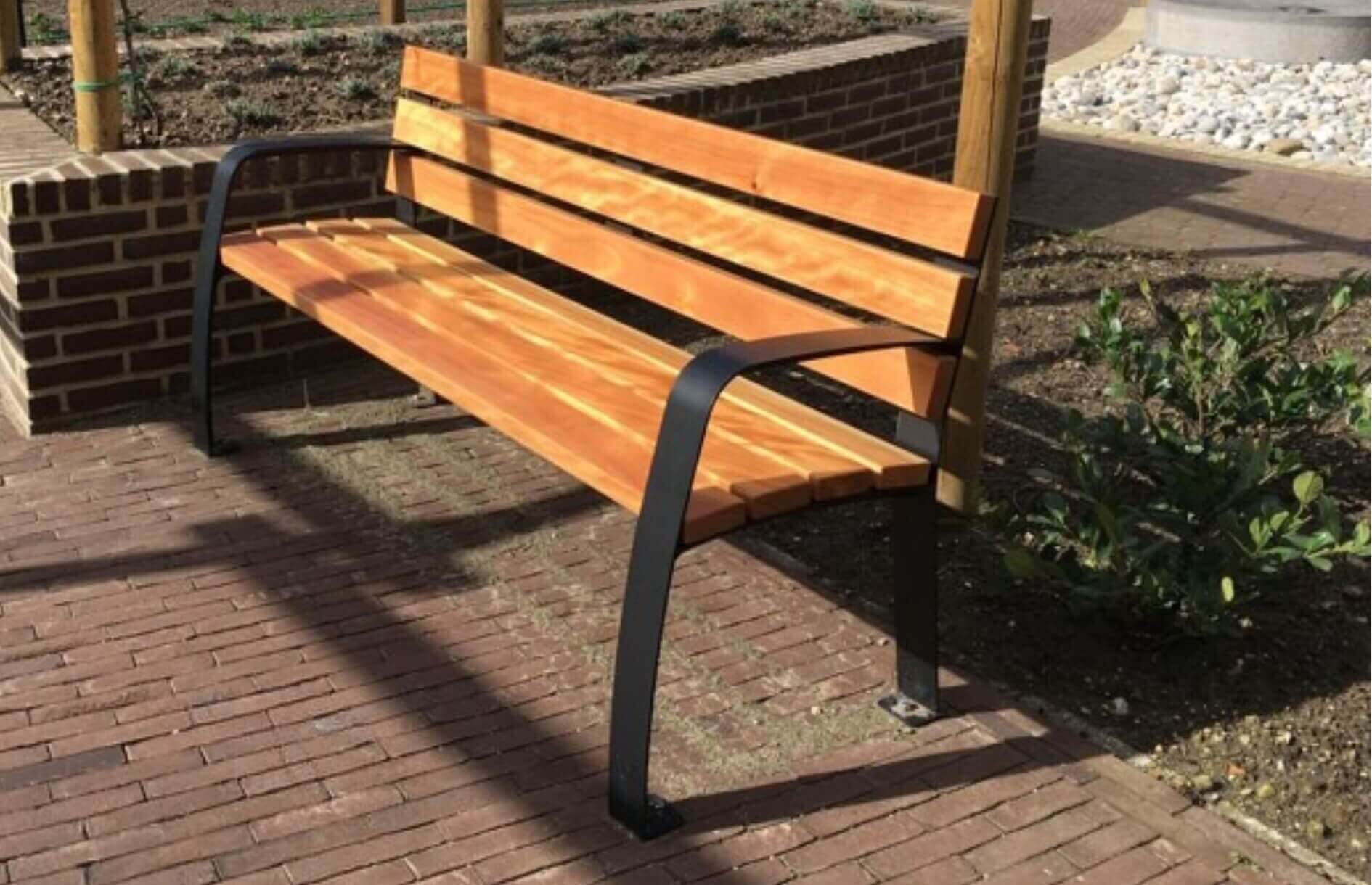 Houten parkbank met ergonomische rugleuning en zwarte stalen armleuningen, geplaatst op een betegeld terras in een verzorgde tuin.