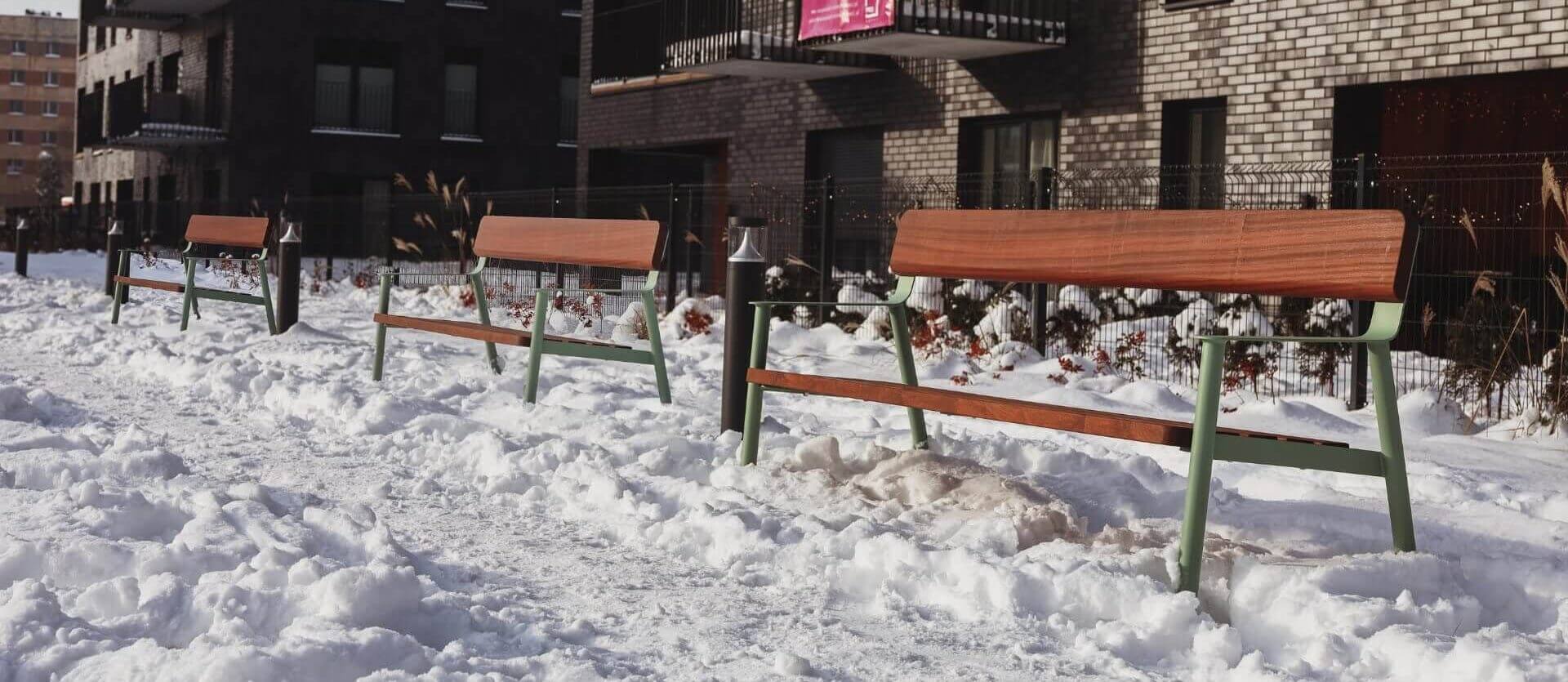 Drie houten parkbanken met groene metalen poten staan in een besneeuwde buitenruimte voor een modern appartementengebouw met balkons.