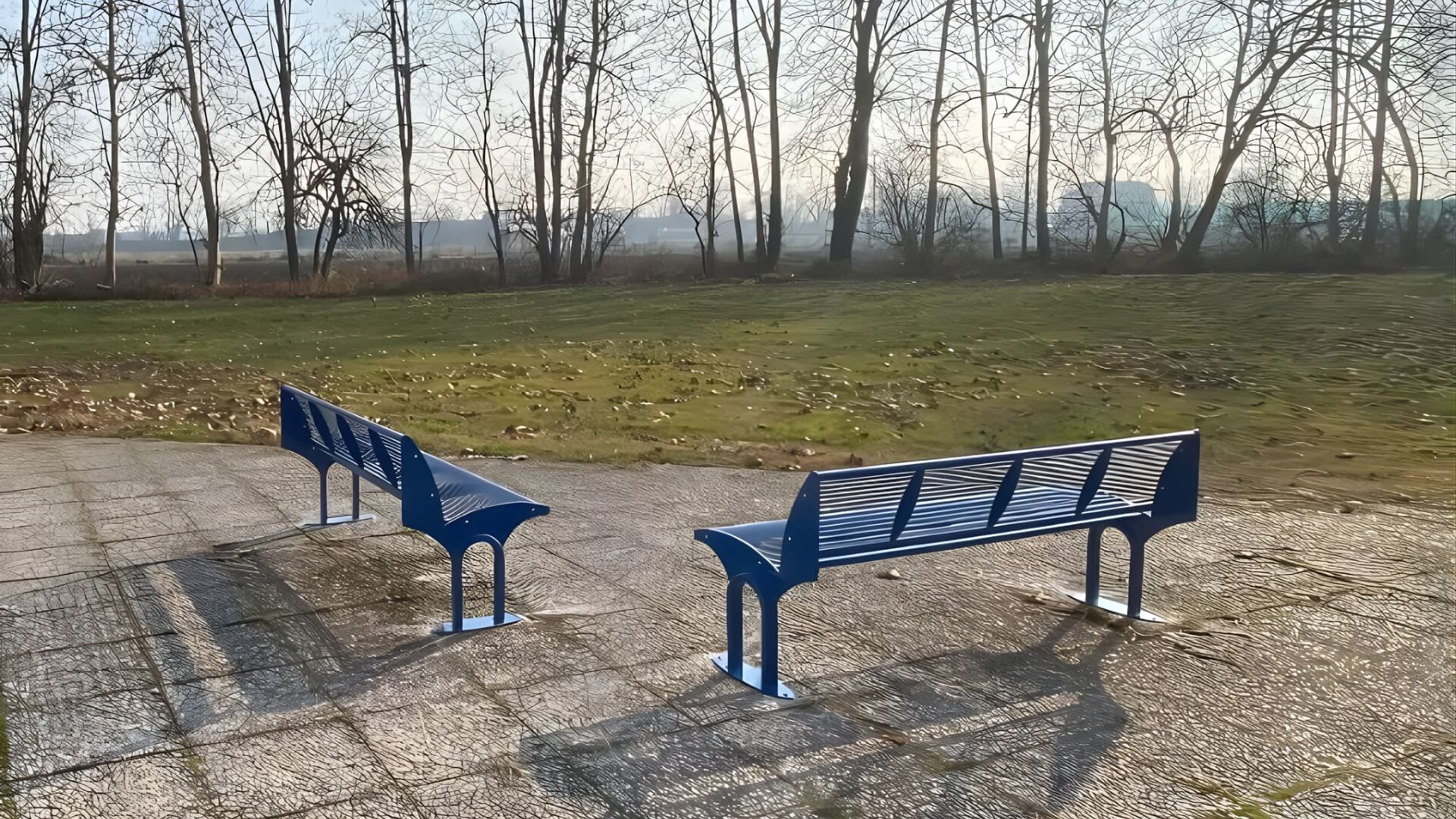 Twee moderne blauwe metalen parkbanken met rugleuning op een betegeld plein met uitzicht op een grasveld en kale bomen in de achtergrond.