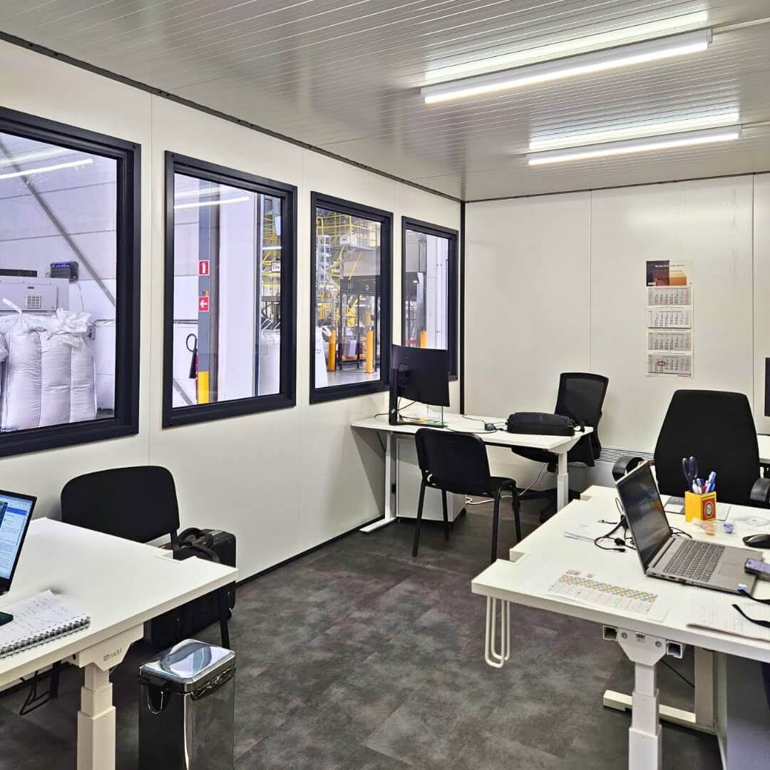 Interieur van een modulaire kantoorunit met bureaus, stoelen, computers en zicht op de productiehal.