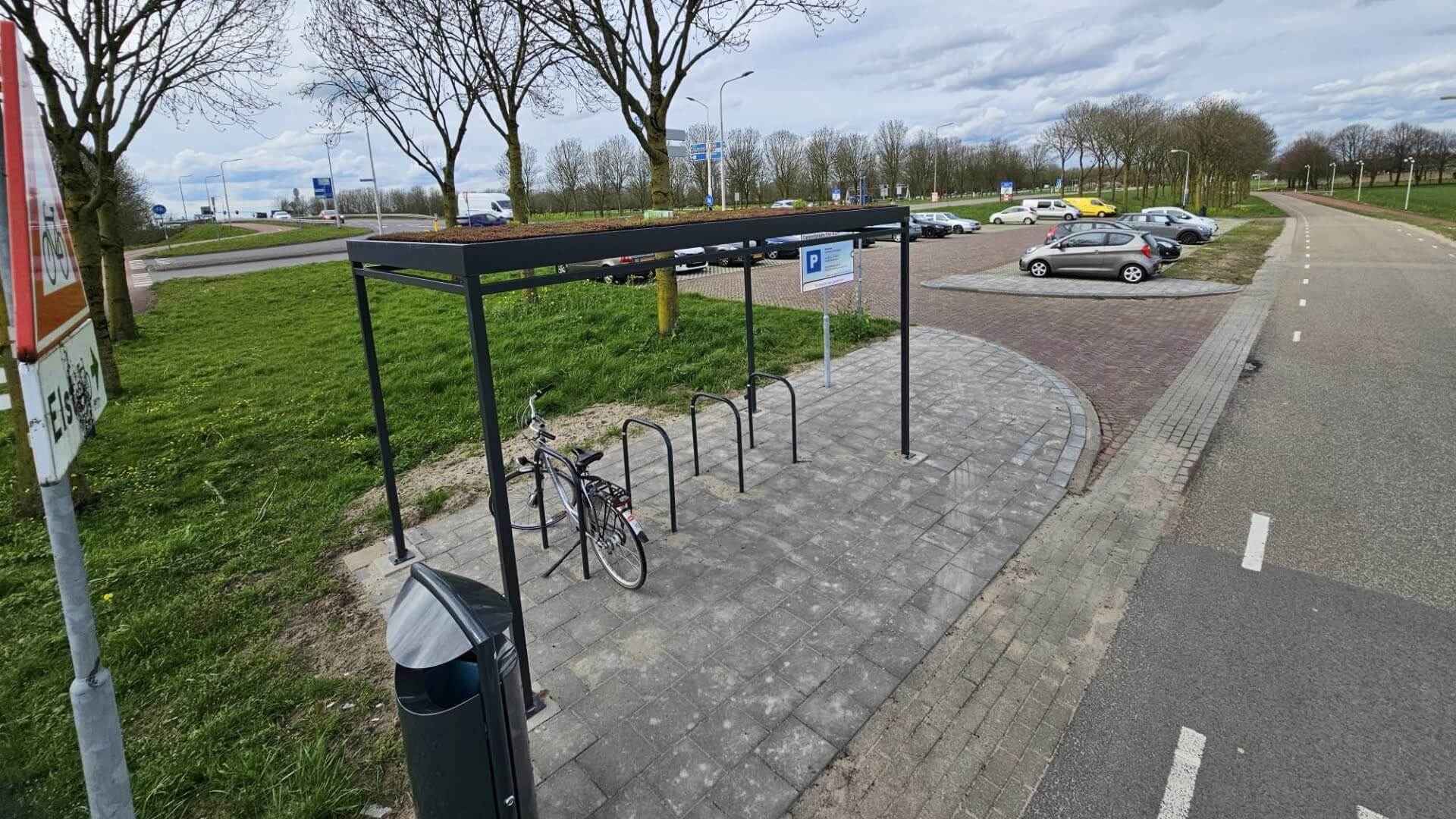 Fietsenstalling met stalen aanleunbeugels onder een moderne fietsoverkapping, geplaatst langs een parkeerterrein en fietspad.
