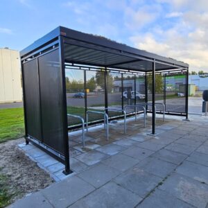 Rijwieloverkapping met vaste fietsenrekken en geïntegreerde oplaadpunten voor elektrische fietsen. Onder de metalen overkapping staan verschillende fietsen gestald, waaronder e-bikes en een scooter.