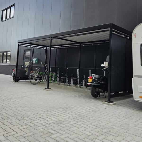 Rijwieloverkapping 6x2m met oplaadpunten voor E-Bike kopen Zwarte overdekte rijwieloverkapping met geïntegreerde fietsenrekken en deels bezet door fietsen, een scooter en een elektrische bakfiets. De constructie staat tegen een modern bedrijfsgebouw met grijze gevelbekleding.
