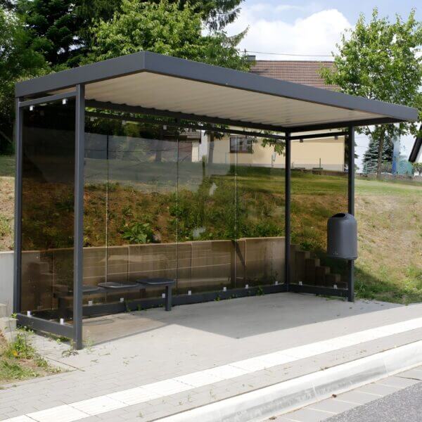 Rookabri bushokje 4x2 meter ral-kleur aanschaffen Donkergrijze rookoverkapping met glazen wanden, ingezet als bushokje langs een weg in een woonomgeving.